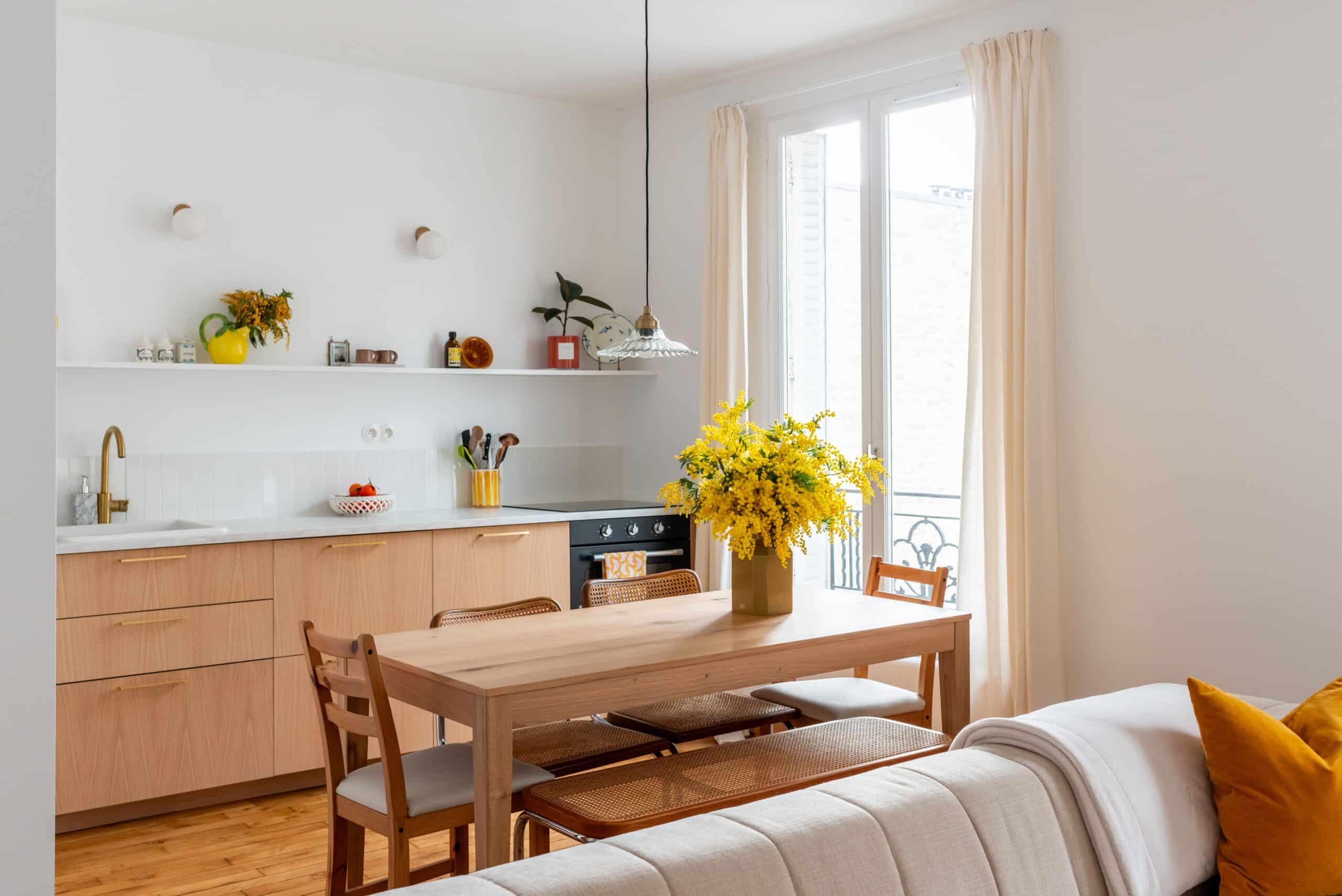 Cette image montre la cuisine d'un appartement parisien, avec une salle à manger lumineuse et aérée, une table et deux chaises. La table a un design simple et moderne aux lignes épurées. Les deux chaises sont rembourrées dans un tissu de couleur claire et ont des dossiers hauts. Un centre de table composé de fleurs colorées trône sur la table.
